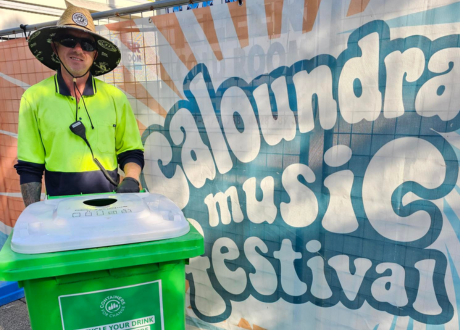 Man in high-vis shirt standing next to a Containers for Change bin in front of a music festival sign