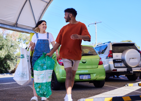 A woman with brown hair, wearing denim overalls and a white shirt is holding a large white bag filled with empty drink containers. A man in a red shirt is beside her, holding a green mesh bag full of empty drink containers.